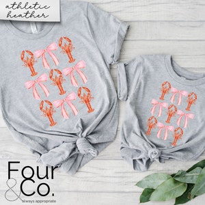 May include: Two grey t-shirts with a pattern of pink bows and orange lobsters. The larger shirt has the text "athletic heather" on the top and "Four & Co. always appropriate" on the bottom.