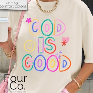 以下が含まれることがあります： クリーム色のTシャツ。カラフルで遊び心のある文字で「GOD IS GOOD」と書かれています。デザインにはスマイリーフェイス、ハート、星が含まれています。このシャツは半袖で、ゆったりとしたフィット感です。