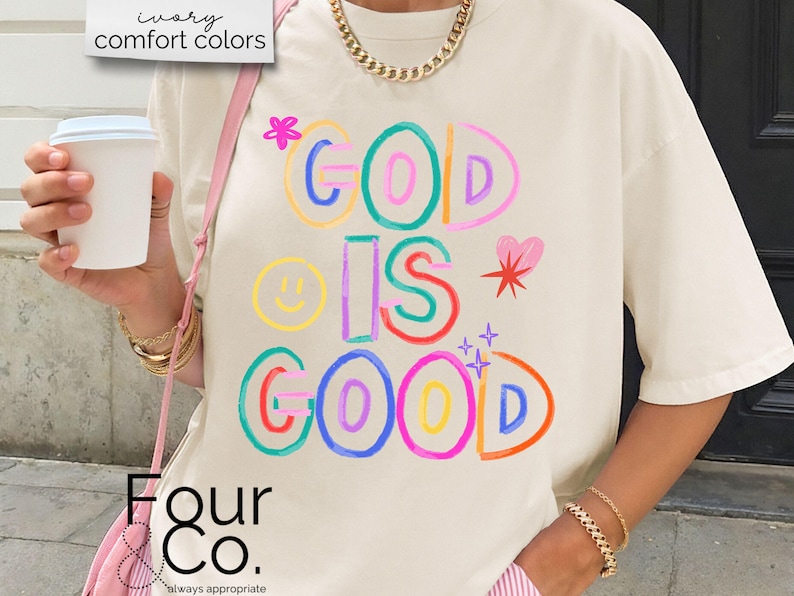 以下が含まれることがあります： クリーム色のTシャツ。カラフルで遊び心のある文字で「GOD IS GOOD」と書かれています。デザインにはスマイリーフェイス、ハート、星が含まれています。このシャツは半袖で、ゆったりとしたフィット感です。