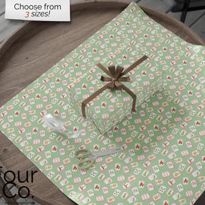 May include: A wrapped gift with a brown bow sits on green wrapping paper patterned with red, white, and orange symbols. A pair of scissors and a roll of ribbon are also on the wrapping paper. The text "Choose from 3 sizes!" is in the top left corner.