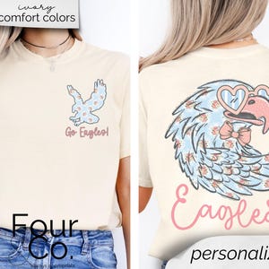 Pu&ograve; includere: T-shirt color crema con un design a forma di aquila. La parte anteriore mostra una piccola grafica di aquila con la scritta "Go Eagles!". Il retro presenta un'aquila pi&ugrave; grande con occhiali a forma di cuore e un papillon, con la parola "Eagles" sotto. La maglietta ha il testo "ivory comfort colors" in alto.