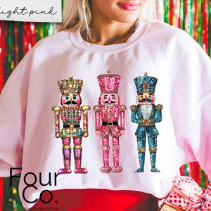 May include: A light pink sweatshirt featuring a design of three jeweled nutcracker figurines. The nutcrackers are wearing traditional costumes and have gold accents. The sweatshirt has the text "Four & Co." printed on the bottom.