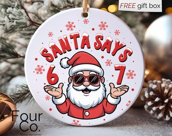May include: A white Christmas ornament with a cartoon Santa Claus wearing sunglasses. The ornament reads "SANTA SAYS 6 7" in red, with snowflakes. A gold string is attached for hanging.