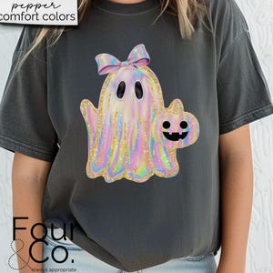 May include: A dark gray t-shirt with a holographic iridescent ghost holding a pumpkin. The ghost has a pink bow on its head. The text "pepper comfort colors" is in the top left corner of the image.