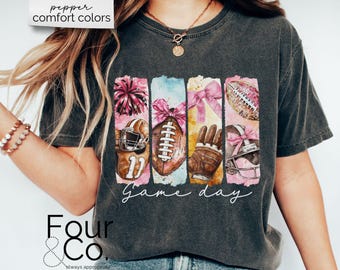 May include: Dark gray t-shirt with a colorful graphic of football-themed elements. The design includes a football helmet, football, and a football glove, with the words "Game day" in a script font. The shirt also has the text "pepper comfort colors" and "Four & Co."