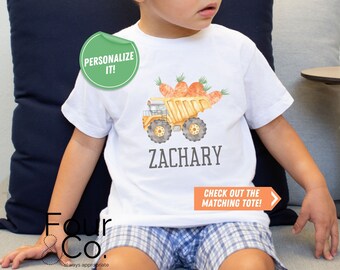 May include: A white t-shirt featuring a yellow toy truck filled with orange carrots. The shirt has the name "ZACHARY" printed on it and the text "CHECK OUT THE MATCHING TOTE!" printed below. The shirt is being worn by a child.