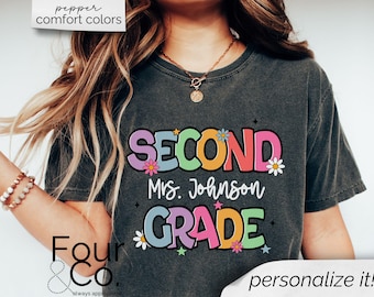 May include: A dark gray t-shirt with a colorful graphic design that says "Second Grade" with the teacher's name "Mrs. Johnson" underneath. The graphic features flowers and stars.