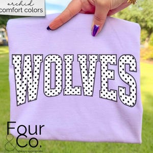 May include: A light purple sweatshirt with the word "WOLVES" in large, black, polka-dotted letters. The sweatshirt also has the text "Four & Co." and "always appropriate" in the bottom left corner. The words "orchid comfort colors" are in the top left corner.