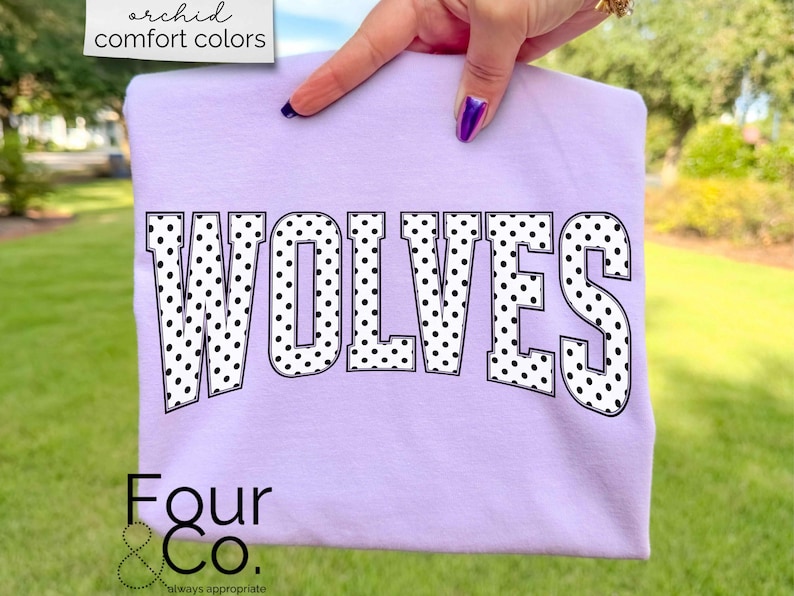 May include: A light purple sweatshirt with the word "WOLVES" in large, black, polka-dotted letters. The sweatshirt also has the text "Four & Co." and "always appropriate" in the bottom left corner. The words "orchid comfort colors" are in the top left corner.