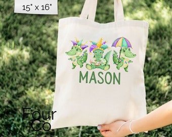May include: A cream-colored tote bag, approximately 15 x 16 inches, featuring three cartoon alligators in Mardi Gras attire. The alligators are green with purple, gold, and green accents. The name "MASON" is printed in green below the alligators.