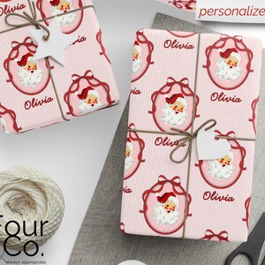 May include: Two wrapped gifts with pink striped paper featuring a Santa Claus design and the name "Olivia." Each package is tied with twine and a white heart-shaped tag. The words "personalize it" are visible.