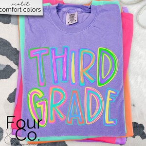 K&ouml;nnte beinhalten: Ein Stapel bunter T-Shirts, darunter ein lila Shirt mit den Worten "THIRD GRADE" in einer lebendigen, mehrfarbigen Schrift. Die Shirts sind auf einer wei&szlig;-schwarz gemusterten Oberfl&auml;che gestapelt. Das oberste Shirt hat ein Etikett mit der Aufschrift "comfort colors".