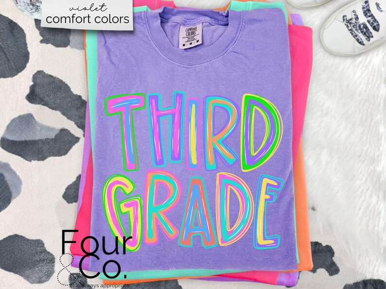 K&ouml;nnte beinhalten: Ein Stapel bunter T-Shirts, darunter ein lila Shirt mit den Worten "THIRD GRADE" in einer lebendigen, mehrfarbigen Schrift. Die Shirts sind auf einer wei&szlig;-schwarz gemusterten Oberfl&auml;che gestapelt. Das oberste Shirt hat ein Etikett mit der Aufschrift "comfort colors".