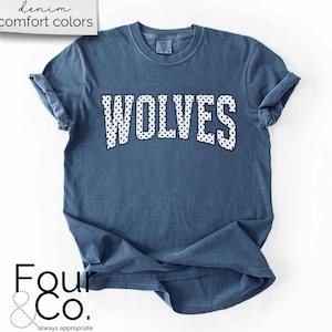 May include: A denim blue t-shirt with the word "WOLVES" in white, polka-dotted letters across the chest. The shirt has rolled-up sleeves and the text "denim comfort colors" is in the upper left corner.
