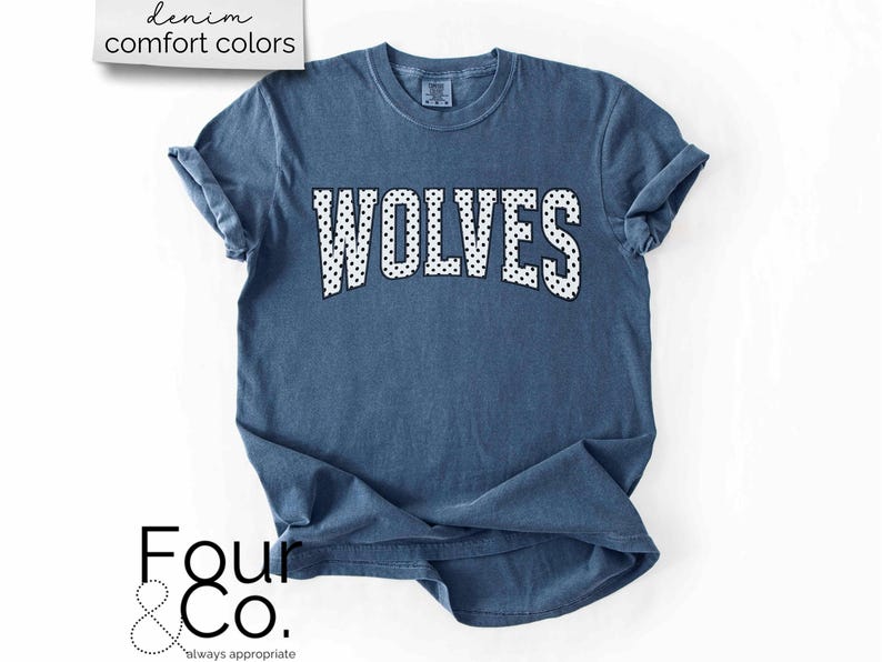 May include: A denim blue t-shirt with the word "WOLVES" in white, polka-dotted letters across the chest. The shirt has rolled-up sleeves and the text "denim comfort colors" is in the upper left corner.