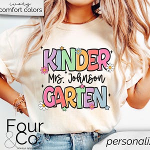 May include: A white t-shirt with the text "Kinder" in rainbow colors, "Mrs. Johnson" in black text, and "Garten" in rainbow colors. The text is surrounded by small flowers and stars. The shirt is being worn by a person with long blonde hair.