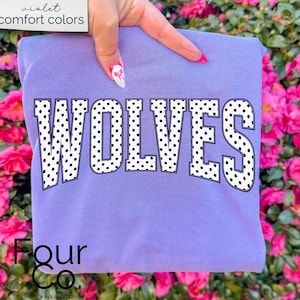 May include: A lilac-colored sweatshirt with the word "WOLVES" in large, white, polka-dotted letters. The text "violet comfort colors" is in the upper left corner. The sweatshirt is held in front of a backdrop of pink flowers.