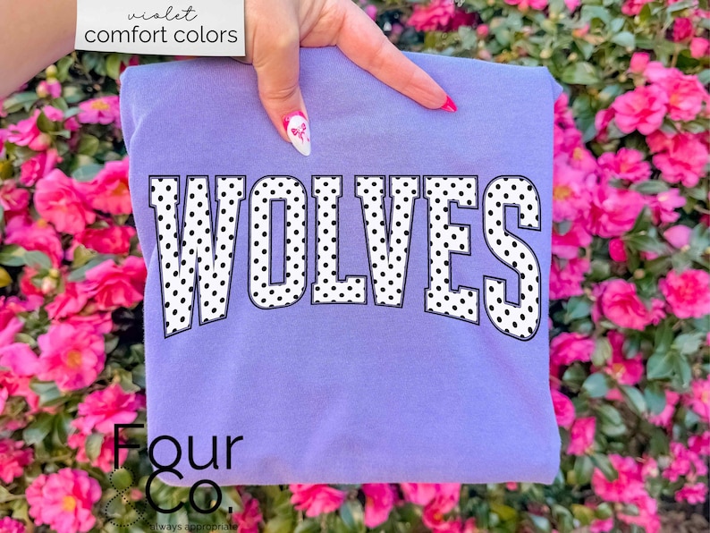 May include: A lilac-colored sweatshirt with the word "WOLVES" in large, white, polka-dotted letters. The text "violet comfort colors" is in the upper left corner. The sweatshirt is held in front of a backdrop of pink flowers.