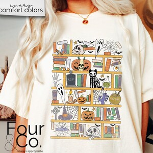 May include: A white t-shirt with a graphic of a bookshelf filled with Halloween-themed items, including books, pumpkins, a ghost, a skeleton cat, and a cauldron. The graphic is in a cartoon style and features a variety of colors, including orange, black, and yellow.