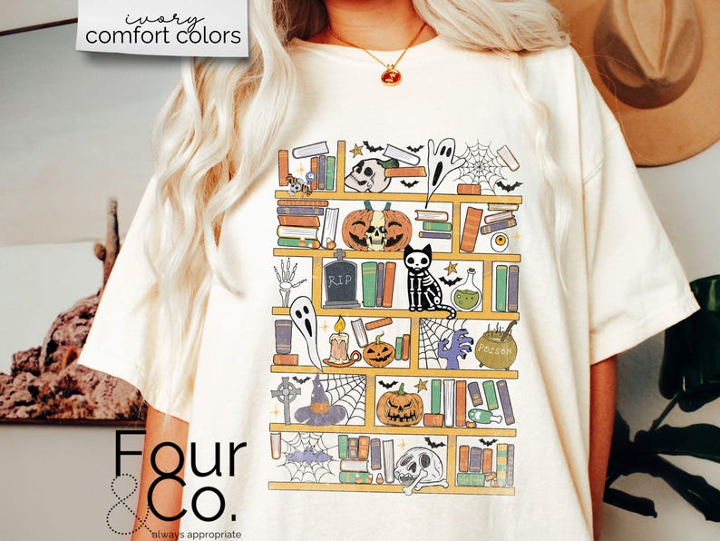 May include: A white t-shirt with a graphic of a bookshelf filled with Halloween-themed items, including books, pumpkins, a ghost, a skeleton cat, and a cauldron. The graphic is in a cartoon style and features a variety of colors, including orange, black, and yellow.