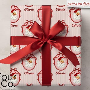 May include: A wrapped gift with a red ribbon and bow. The wrapping paper features a repeating pattern of Santa Claus faces in pink frames with the name "Olivia" and a striped background. The words "personalize it" are in the top right corner.