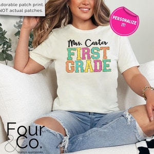 May include: A white t-shirt with a colorful "Mrs. Carter First Grade" design. The text is in a retro font with a glittery effect. The shirt is being worn by a person sitting on a white couch.