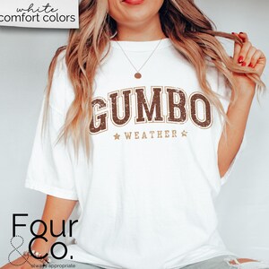 May include: White t-shirt with a brown and gold graphic that says "GUMBO WEATHER" with stars on either side of the text.