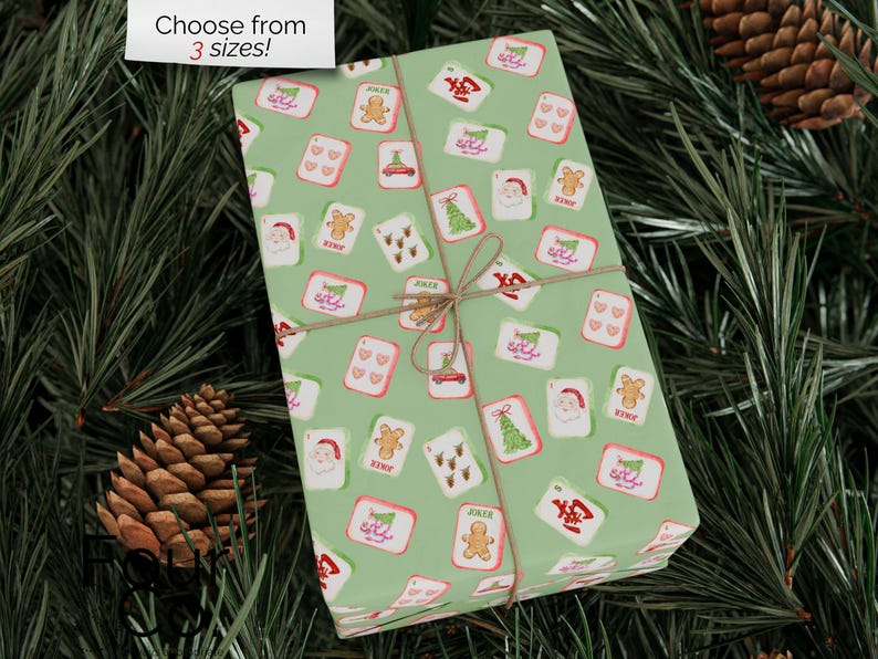 May include: A wrapped gift with a light green background, adorned with a repeating pattern of Christmas-themed playing cards. The cards feature images of Santa, gingerbread men, Christmas trees, and other holiday symbols. The gift is tied with twine and the text "Choose from 3 sizes!" is in the top left corner.