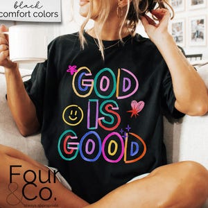 以下が含まれることがあります： カラフルなアウトライン文字で「GOD IS GOOD」と書かれた黒いTシャツ。デザインにはスマイリーフェイス、ハート、星が含まれています。「black comfort colors」という言葉が左上の白いボックスにあります。