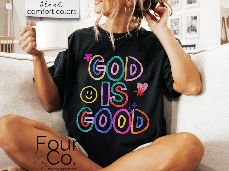 以下が含まれることがあります： カラフルなアウトライン文字で「GOD IS GOOD」と書かれた黒いTシャツ。デザインにはスマイリーフェイス、ハート、星が含まれています。「black comfort colors」という言葉が左上の白いボックスにあります。