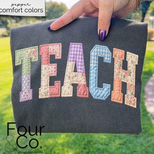 Peut inclure: Un sweat-shirt gris foncé avec le mot "TEACH" appliqué en tissu patchwork coloré. Les lettres sont de style universitaire, avec des motifs floraux, vichy et unis. Le texte "pepper comfort colors" est en haut à gauche.