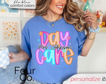 May include: A light blue t-shirt with the words "Day Care Mrs. Johnson" in a colorful, playful font. The shirt has a relaxed fit and is suitable for casual wear. The text "personalize it!" is also visible.