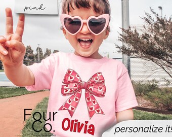 May include: Pink t-shirt with a red and white checkered bow and the text "Four & Co. Olivia" printed on the front.