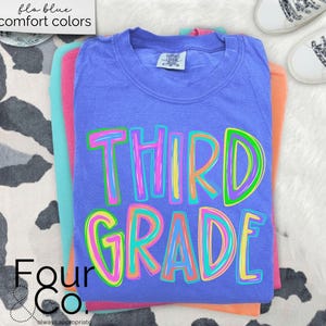 K&ouml;nnte beinhalten: Ein Stapel bunter T-Shirts, wobei das blaue Shirt oben die Worte "THIRD GRADE" in einer lebendigen, mehrfarbigen Schrift tr&auml;gt. Die Shirts sind auf einer wei&szlig;en, strukturierten Oberfl&auml;che gestapelt, mit einem Paar wei&szlig;er Turnschuhe im Hintergrund. Der Text "fla blue comfort colors" befindet sich oben links.
