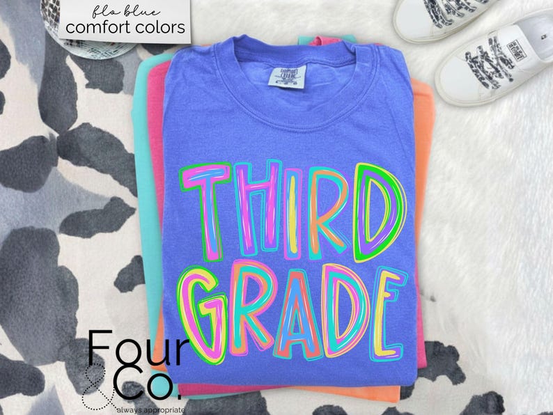 K&ouml;nnte beinhalten: Ein Stapel bunter T-Shirts, wobei das blaue Shirt oben die Worte "THIRD GRADE" in einer lebendigen, mehrfarbigen Schrift tr&auml;gt. Die Shirts sind auf einer wei&szlig;en, strukturierten Oberfl&auml;che gestapelt, mit einem Paar wei&szlig;er Turnschuhe im Hintergrund. Der Text "fla blue comfort colors" befindet sich oben links.