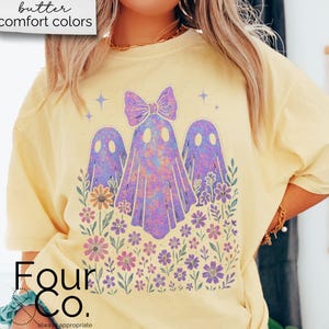 Puede incluir: Camiseta amarilla clara con un gr&aacute;fico de tres fantasmas morados con un lazo, rodeados de flores. El texto "butter comfort colors" est&aacute; en la esquina superior izquierda. La marca "Four Co." est&aacute; en la parte inferior.