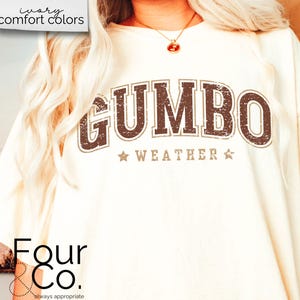 May include: A white t-shirt with a brown distressed graphic that says "GUMBO WEATHER" with stars on either side of the text. The text is in a vintage style font. The shirt is from Four & Co. and says "always appropriate" below the logo.