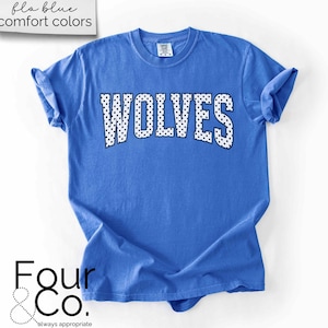 May include: A blue t-shirt with the word "WOLVES" in white letters with black polka dots. The shirt has rolled-up sleeves and the text "flo blue comfort colors" is in the upper left corner. The logo "Four & Co." is in the lower left corner.