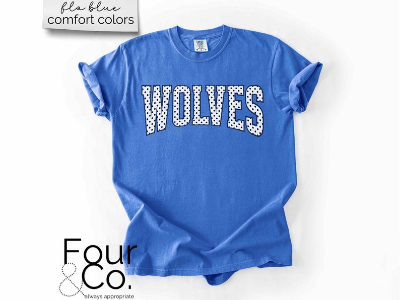 May include: A blue t-shirt with the word "WOLVES" in white letters with black polka dots. The shirt has rolled-up sleeves and the text "flo blue comfort colors" is in the upper left corner. The logo "Four & Co." is in the lower left corner.
