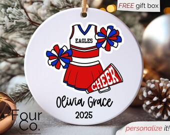 May include: White round ornament featuring a cheerleader uniform design in red, white, and blue. The ornament includes the text "EAGLES", "CHEER", "Olivia Grace", and "2025". A gold string is attached for hanging.