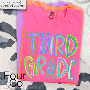 K&ouml;nnte beinhalten: Ein Stapel T-Shirts in Neonrosa, Orange und Lila. Das vordere Shirt ist leuchtend rosa mit den Worten "THIRD GRADE" in bunten, umrandeten Buchstaben. Die Shirts sind mit "neon pink comfort colors" beschriftet. In der unteren linken Ecke steht der Text "Four Co."