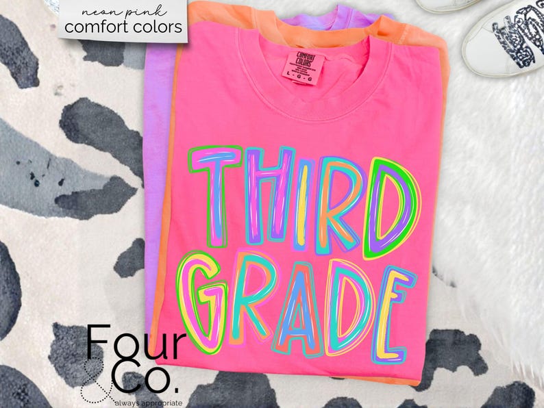 K&ouml;nnte beinhalten: Ein Stapel T-Shirts in Neonrosa, Orange und Lila. Das vordere Shirt ist leuchtend rosa mit den Worten "THIRD GRADE" in bunten, umrandeten Buchstaben. Die Shirts sind mit "neon pink comfort colors" beschriftet. In der unteren linken Ecke steht der Text "Four Co."