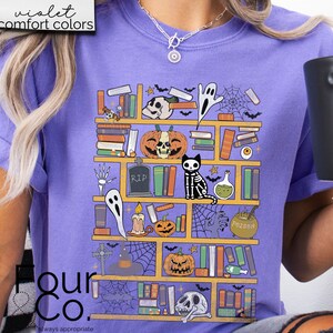 May include: A purple t-shirt with a graphic of a bookshelf filled with books, skulls, ghosts, candles, and a black cat. The text "Poison" is on a potion bottle on the shelf.