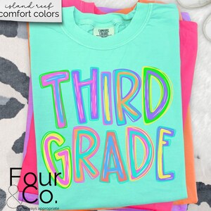 K&ouml;nnte beinhalten: Ein gefaltetes, hellt&uuml;rkisfarbenes T-Shirt mit den Worten "THIRD GRADE" in gro&szlig;en, bunten Buchstaben. Das Shirt geh&ouml;rt zur "island reef comfort colors"-Kollektion und ist auf rosa und orangefarbenen Shirts gestapelt. Das Four & Co. Logo ist sichtbar.