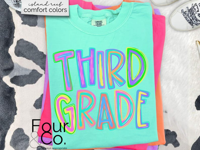 K&ouml;nnte beinhalten: Ein gefaltetes, hellt&uuml;rkisfarbenes T-Shirt mit den Worten "THIRD GRADE" in gro&szlig;en, bunten Buchstaben. Das Shirt geh&ouml;rt zur "island reef comfort colors"-Kollektion und ist auf rosa und orangefarbenen Shirts gestapelt. Das Four & Co. Logo ist sichtbar.