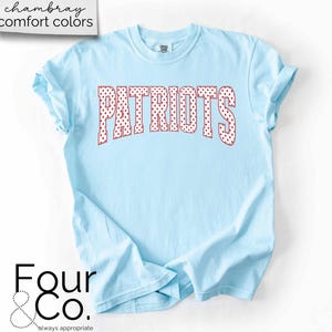 May include: Light blue t-shirt with the word "PATRIOTS" in red polka dot letters. The shirt has rolled-up sleeves and the text "chambray comfort colors" is in the upper left corner. The logo "Four & Co." is in the lower left corner.