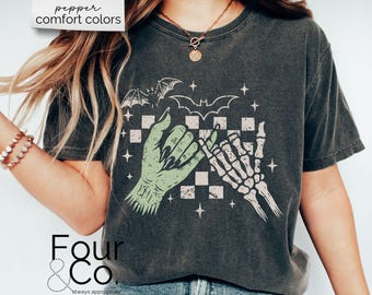 May include: Dark gray t-shirt with a graphic of a green hand and a skeleton hand pinky-promising. The design includes bats, stars, and a checkered background. The text "pepper comfort colors" is at the top.
