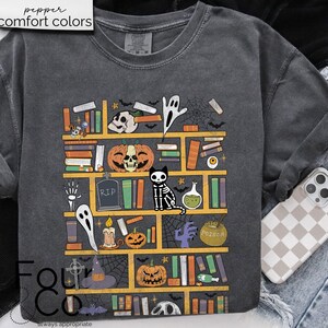 May include: A dark gray t-shirt with a graphic of a bookshelf filled with spooky Halloween themed items. The graphic includes books, pumpkins, ghosts, a skeleton cat, a potion bottle, and a witch's hat. The text "RIP" and "Poison" are visible on the graphic.