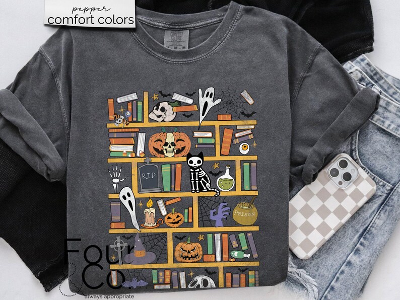 May include: A dark gray t-shirt with a graphic of a bookshelf filled with spooky Halloween themed items. The graphic includes books, pumpkins, ghosts, a skeleton cat, a potion bottle, and a witch's hat. The text "RIP" and "Poison" are visible on the graphic.