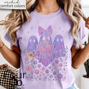 Puede incluir: Camiseta lila claro con un dise&ntilde;o de tres fantasmas con un lazo, rodeados de flores. Los fantasmas tienen un efecto tie-dye colorido. El texto "orchid comfort colors" est&aacute; en la esquina superior izquierda.